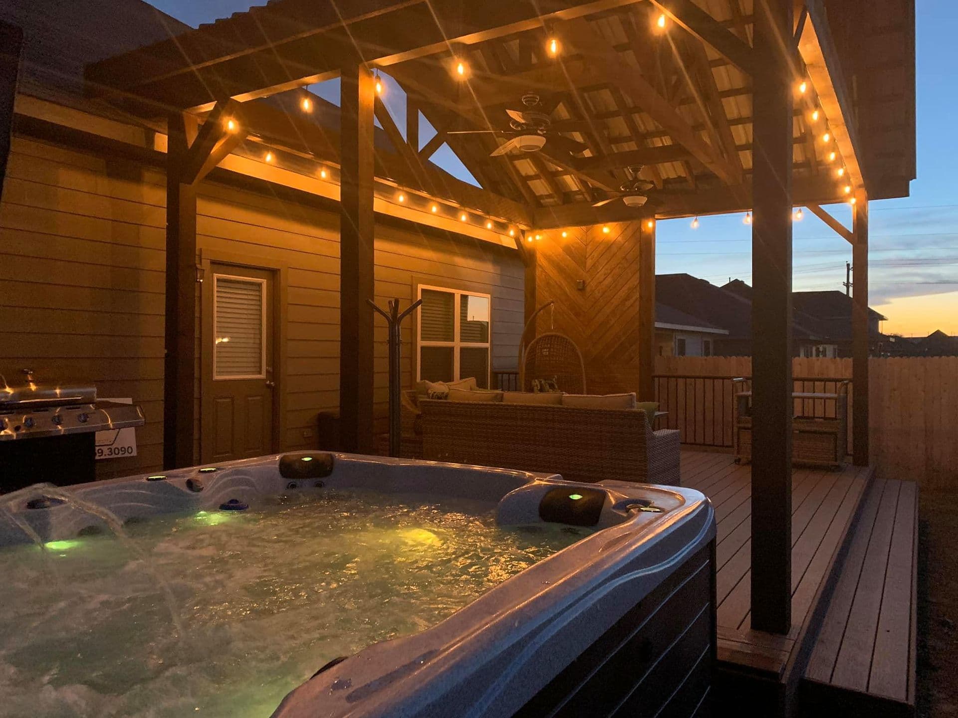 Covered deck at dusk with string lights, ceiling fans, diagonal wood accent wall, hot tub