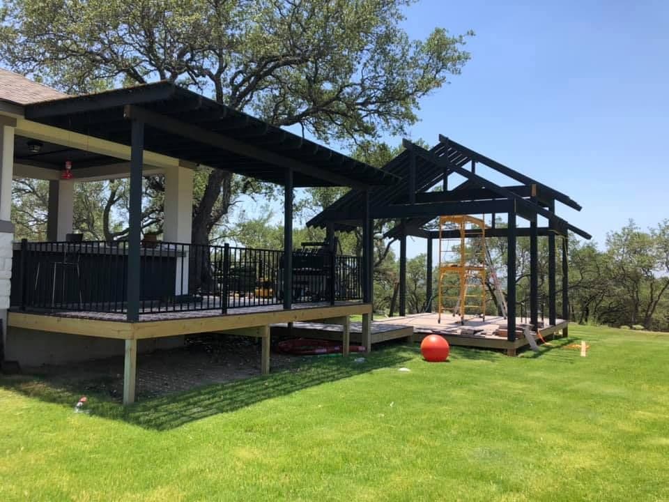 Raised covered deck with black railing beside a gabled pavilion frame among Central Texas oaks