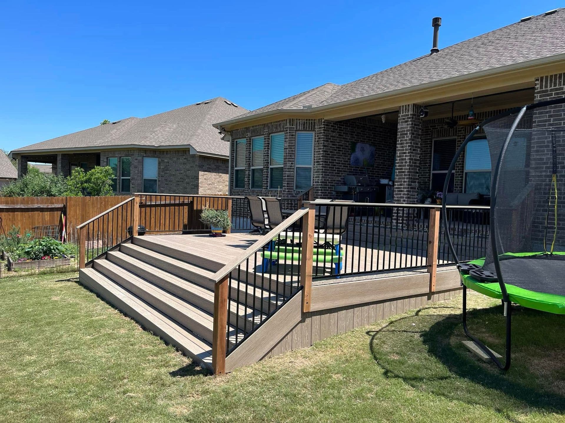 Wide composite deck with cascading stairs, cedar posts, and black metal balusters behind a brick home